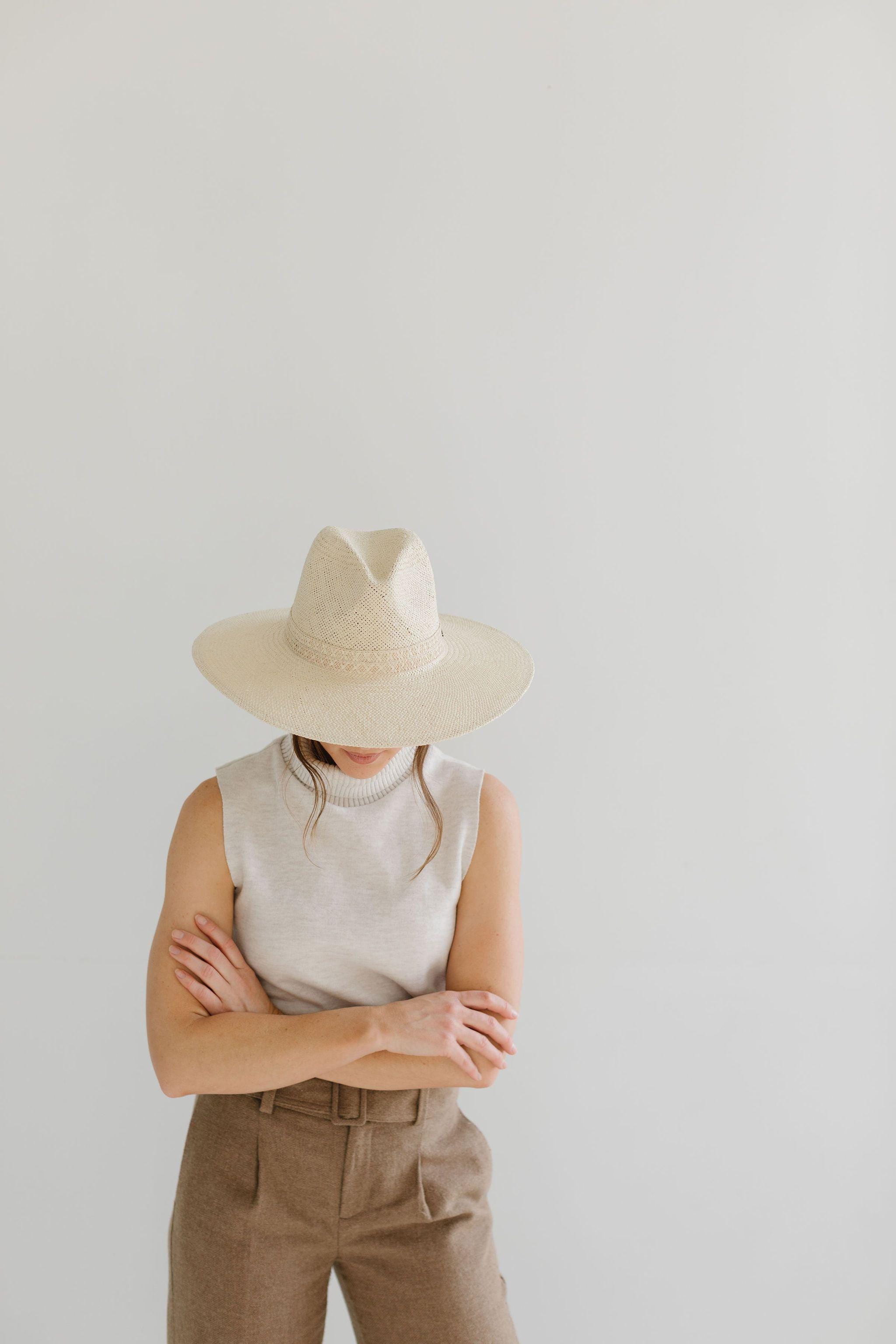 Woman wearing a cream straw hat, a white sleeveless top and brown pants, looking down against a plain background #color_cream