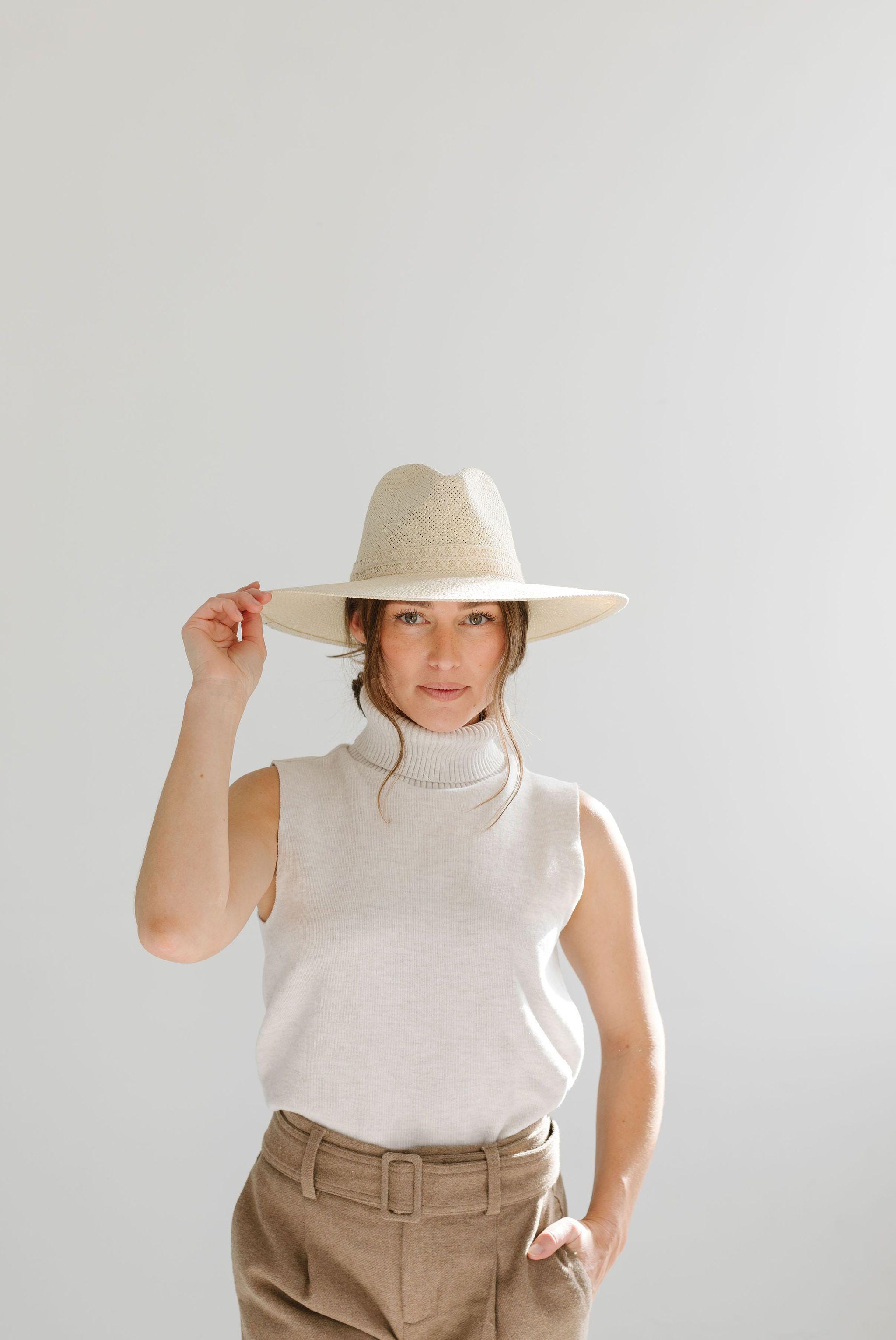 Woman wearing a cream straw hat, a white sleeveless top and brown pants against a plain background #color_cream