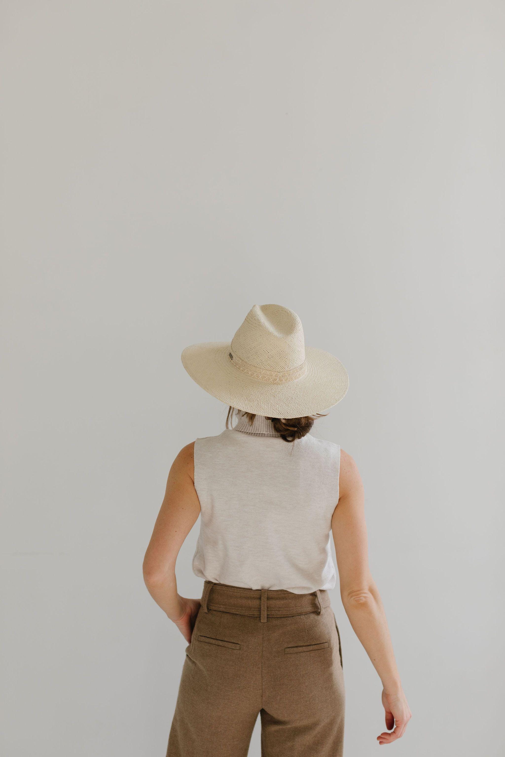 Woman wearing a cream straw hat, a white sleeveless top and brown pants, facing behind against a plain background #color_cream