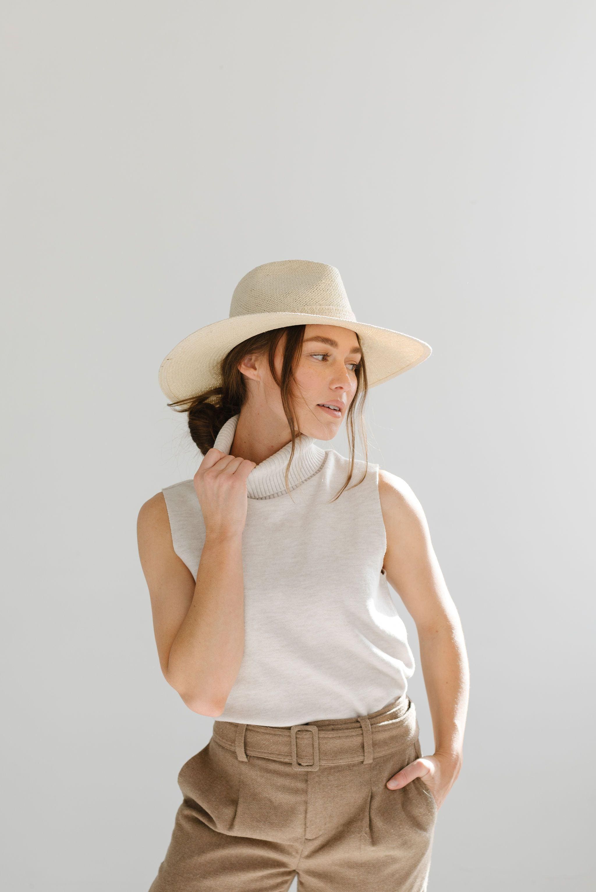 Woman wearing a cream straw hat, a white sleeveless top and brown pants, looking sideways against a plain background #color_cream